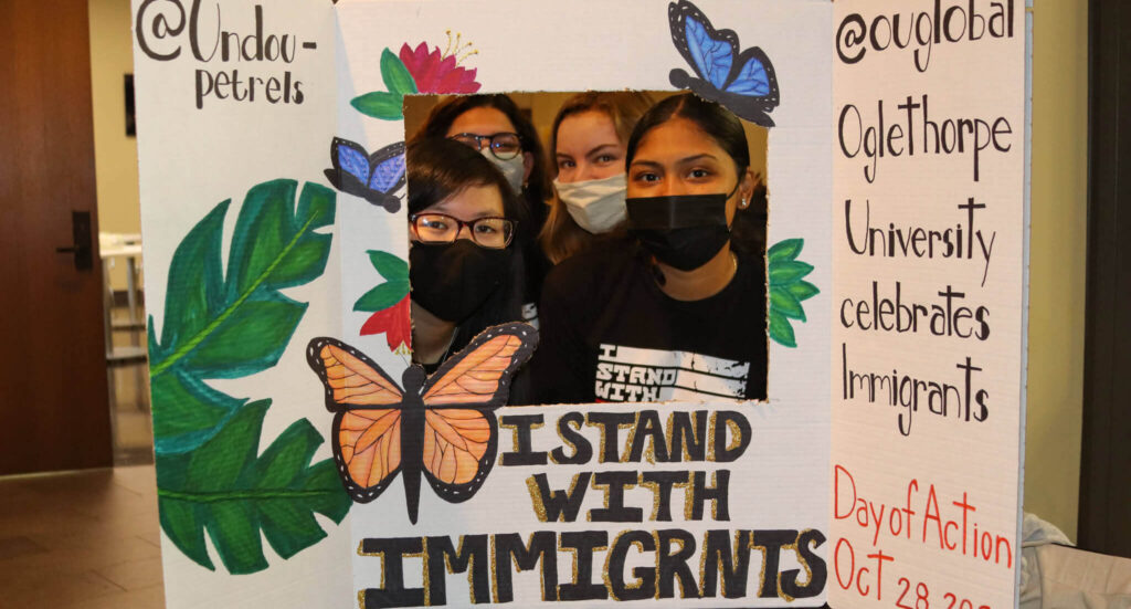 Four students pose inside a poster frame that reads "I stand with immigrants."