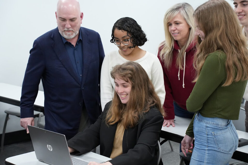 Students and instructors gather around a student and her laptop in a classroom.