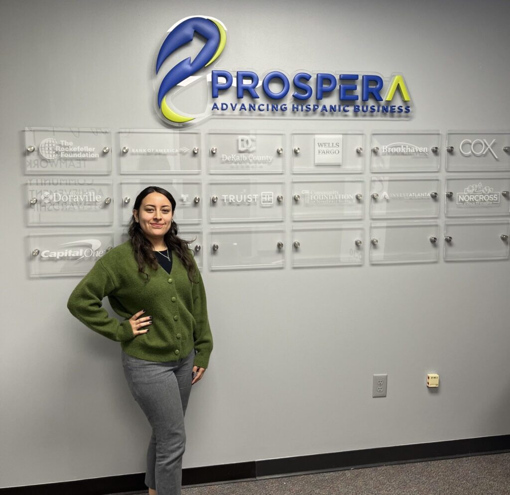 Alexa Miranda at the Prospera office posing in front of a wall with the organization's logo and partners which include Truist, the City of Brookhaven, Capital one, and more.