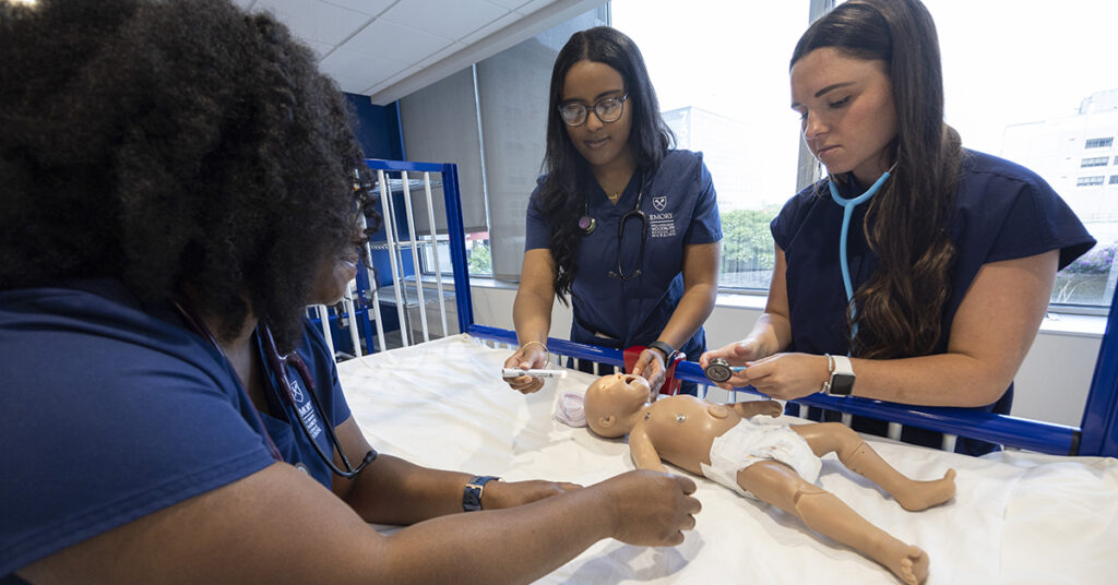 Three nursing students in scrub check vital signs on a mannequin baby