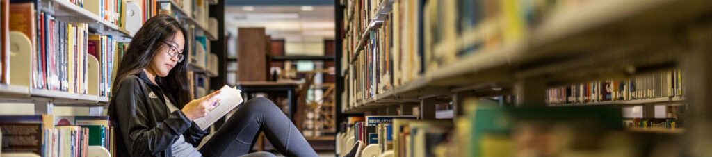 A student reads a book on ground in an aisle of books in the library