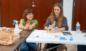 Professor Abigail Gregg watches a student sculpt a figure with clay while they both smile.