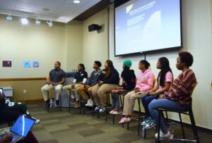 The 8 panel guest speakers pictured sitting alongside the moderator in the front of the room.