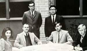 A black and white photo of two male faculty members in suits standing behind three seated male students in coats and ties and a female student in a blazer.
