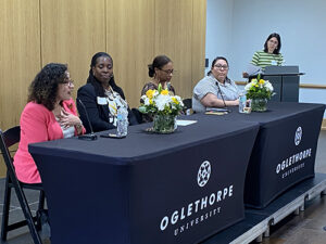 Four women sitting at a table on a dais with a woman at a podium on the right