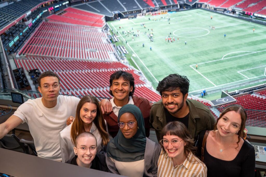Eight students smile for a photo from the press box. A soccer stadium can be seen behind them.