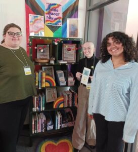 Fynn Grindle, Jennah Waters, and Gabby Atencio pose next to the little queer library that is full with books, resources, and decorations.
