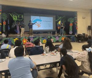 David Raygoza and other students stand on a stage and give a presentation on air resistance to elementary students.