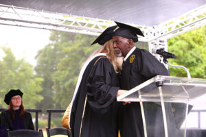 A man in a cap and gown hugs a woman in a cap and gown on stage