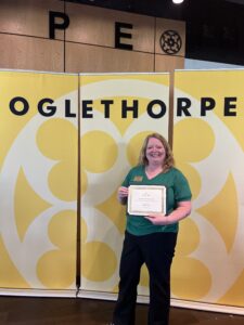 Heather Neal poses with her Excellence in Service award in front of a yellow Oglethorpe backdrop.