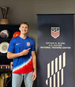 Jack Lowrie wearing a USA soccer jersey pointing next to a sign that says "Arthur M Blankc U.S. Soccer National Training Center."