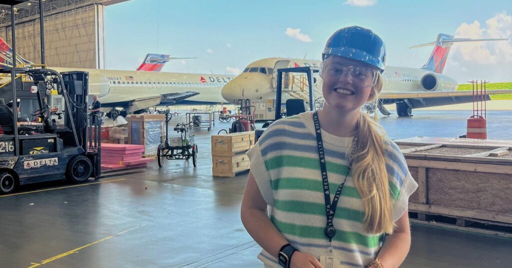 A student stands in a hangar with airplanes and other machinery visible behind her