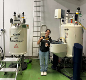 Emily Mendendez wears lab goggles in and smiles in between two Nuclear Magnetic Resonance machines.