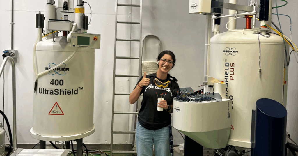 Emily Mendendez wears lab goggles in and smiles in between two Nuclear Magnetic Resonance machines.
