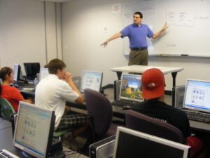 A teacher points to a whiteboard with mathematical equations as students at their computers look on