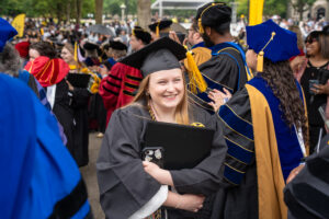 2026 graduate wearing cap and gown smiles brightly holding her degree while walking through a crowd of professors clapping and cheering for graduates.