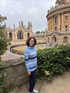 Student wearing a striped sweater standing in front of academic buildings with distinct architecture.