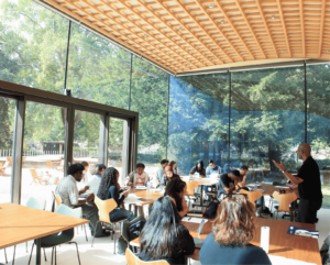 Students gathered in a room with large windows listens to a speaker at a podium.