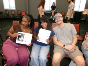 Three students smiling in casual clothes, with two holding up scripts for a play.