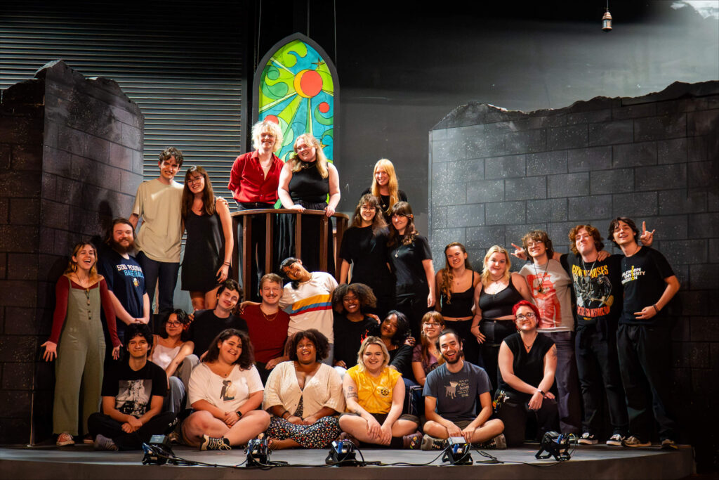 Large group of theatre cast and crew on stage with stylized set, featuring broken walls and a stained glass window.